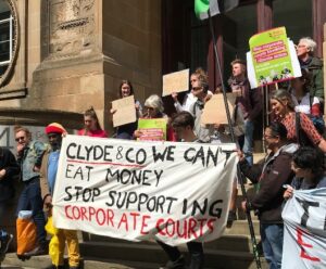 protestors outside lawyers firm Clyde & Co with a banner that reads 'we can't eat money stop supporting corporate courts'